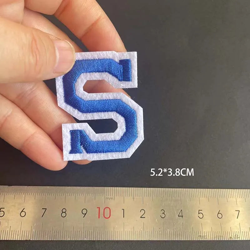 A hand holds a blue alphabet embroidery iron-on patch shaped like the letter S against a ruler.