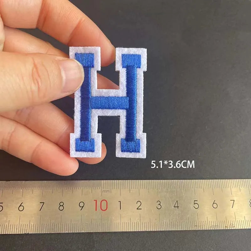 A hand holds a blue embroidery patch shaped like the letter H, measured against a ruler.