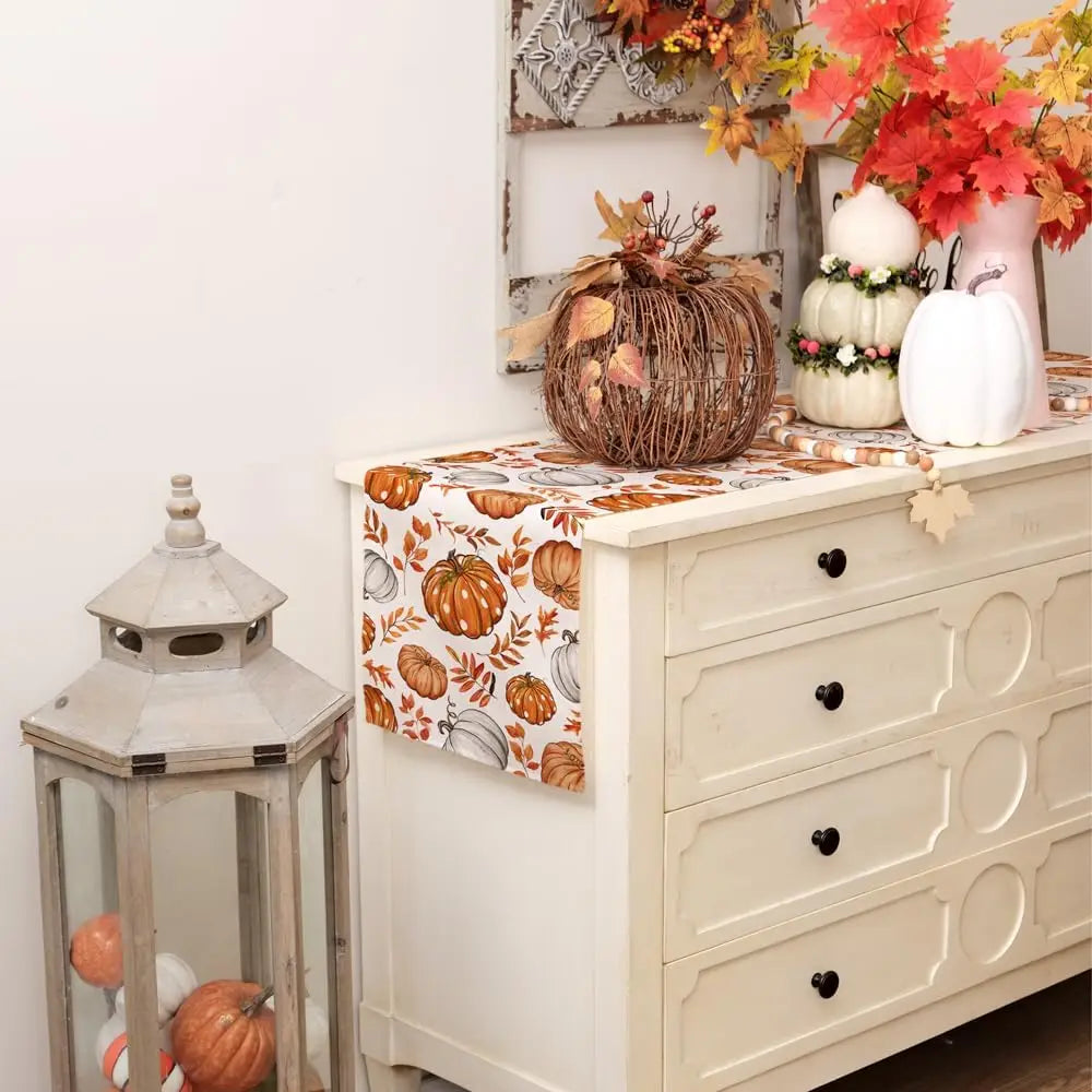 A linen table runner for autumn decor featuring pumpkins and foliage on a white wooden cabinet.