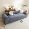 A light blue quilted sofa cover with elastic straps is displayed on a beige rug in a bright living room.