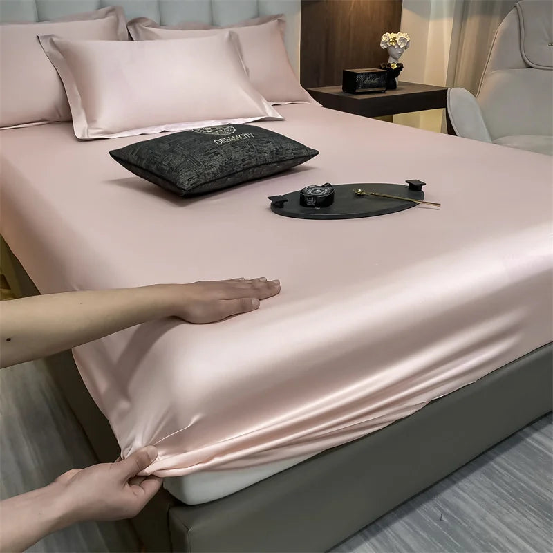 A pink fitted bed sheet is being adjusted on a bed, with pillows and a dark cloth resting on the surface.