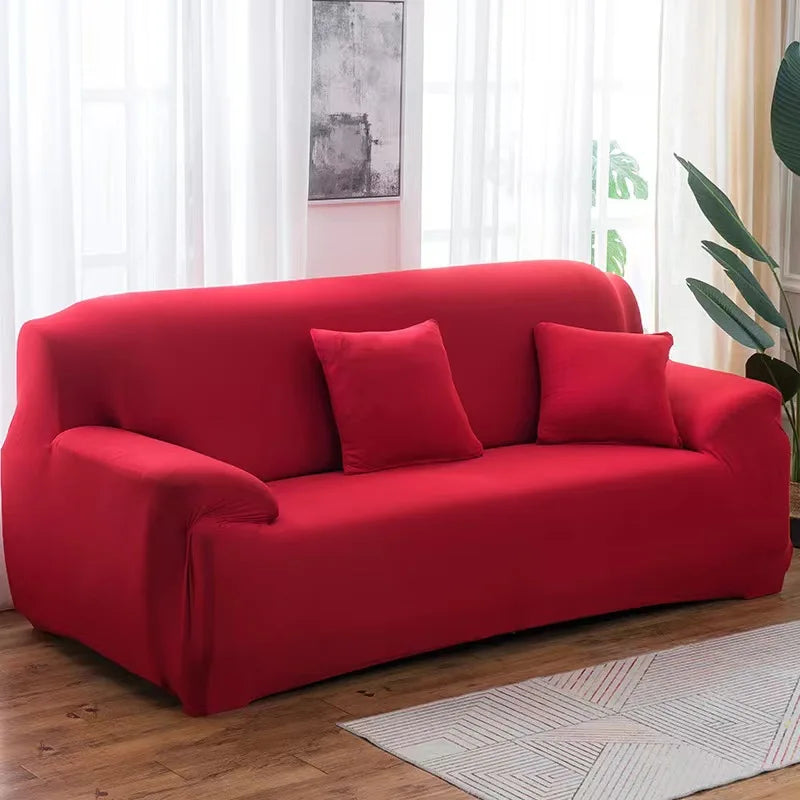 A vibrant red stretch sofa cover with two accent pillows placed on a wooden floor near a window.