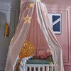 A pink princess hanging bed canopy with sheer fabric and gold string lights, featuring a wooden crib.