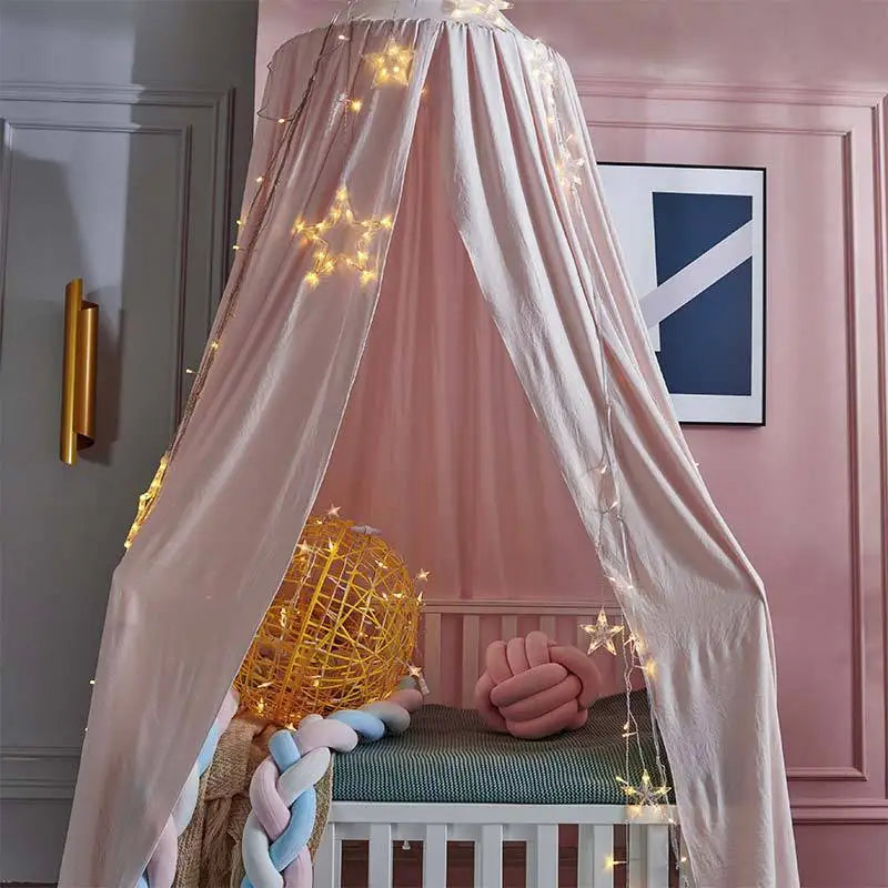 A pink princess hanging bed canopy with sheer fabric and gold string lights, featuring a wooden crib.