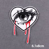 A black and white embroidered heart-shaped patch featuring a stylized, bloody eye design.