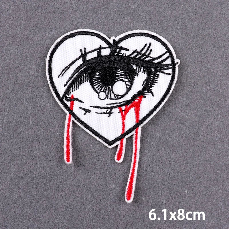 A black and white embroidered heart-shaped patch featuring a stylized, bloody eye design.