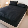 A black quilted mattress protector is shown on a bed, with pillows and a bedside lamp visible.