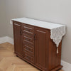 A dark wood cabinet with a white embroidered lace table runner on top, used for dining or furniture accents.