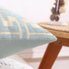 A close-up of a light blue throw pillow cover with beige geometric embroidery resting on a wooden surface.