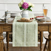 A light green embroidered cotton linen table runner with white tassels placed on a wooden table.