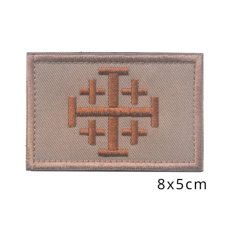A rectangular embroidered patch featuring a stylized cross design in brown thread on a beige fabric background.