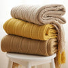 A stack of beige, yellow, and tan knitted tassel throw blankets rolled up on a white stool.
