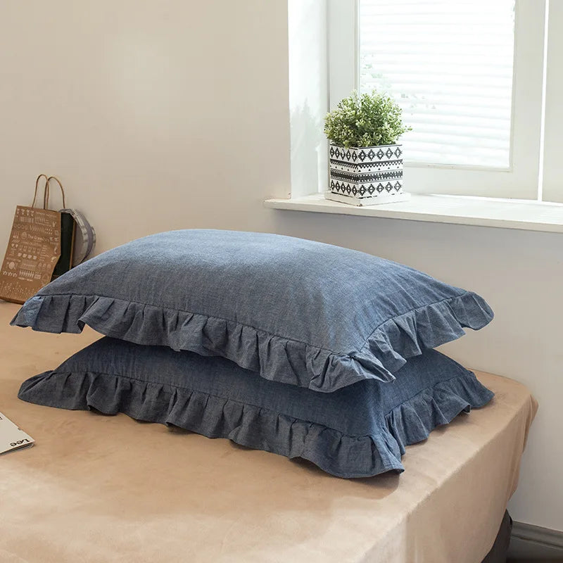 A set of two soft blue washed cotton pillowcases with ruffled edges resting on a light brown surface.