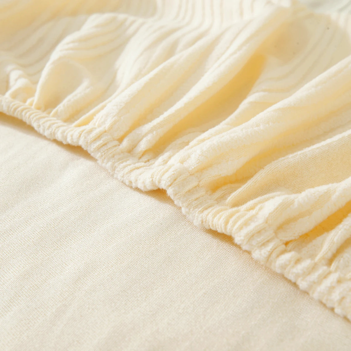 A close-up of a cream-colored stretchable sofa cover with a ribbed line pattern, showing the gathered hemline.
