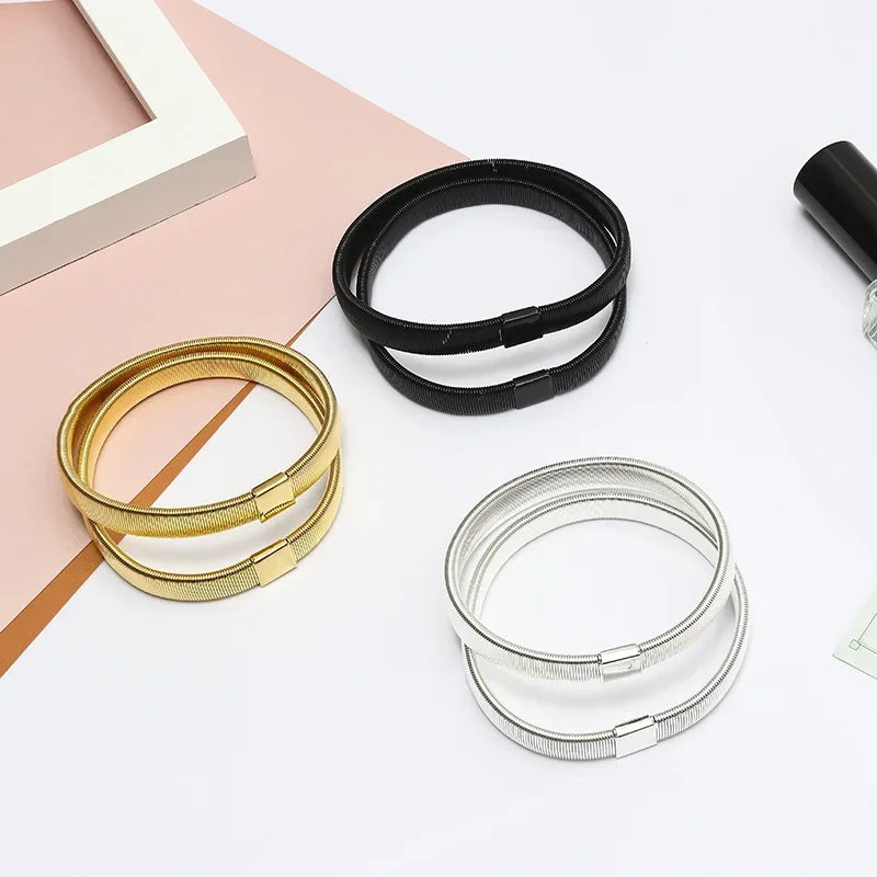 Three adjustable metal sleeve garters for men in gold, black, and silver lying on a pink surface.