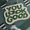 A white foot mat with green embossed text that reads YOU LOOK GOOD placed on a teal rug.