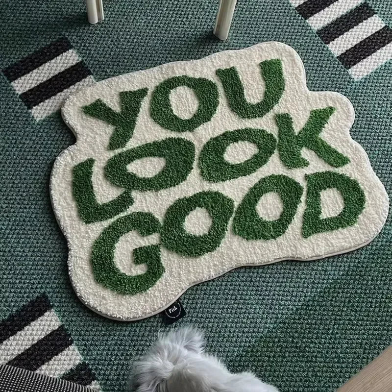A white foot mat with green embossed text that reads YOU LOOK GOOD placed on a teal rug.