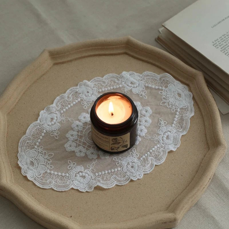 A small candle in a glass jar rests on a white lace doily on a beige ceramic coaster.