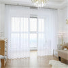 White semi-sheer tulle curtains covering a large window with natural light and wooden flooring.