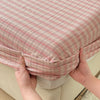 Hands are holding a pink and white plaid fitted sheet on a bed.