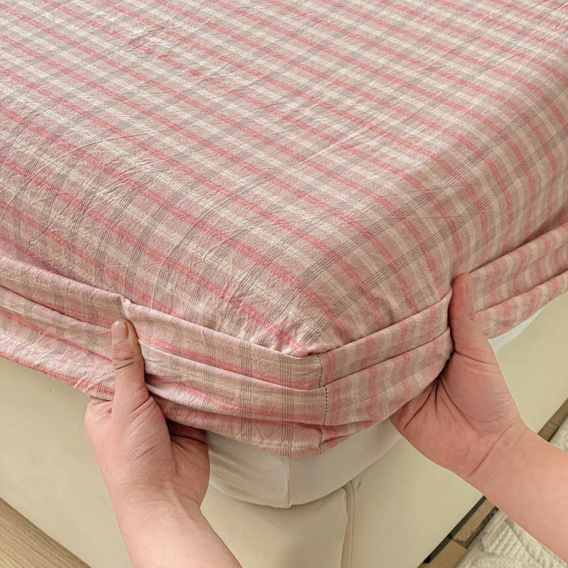 Hands are holding a pink and white plaid fitted sheet on a bed.