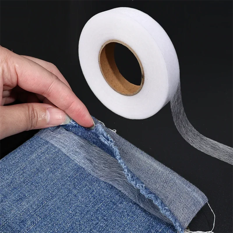 A hand is tearing a piece of denim fabric to show double-sided iron-on adhesive tape for garment repair rolled on a white dis