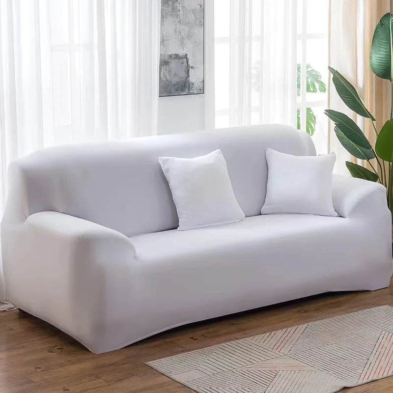 A modern, light gray stretch sofa cover placed in a bright living room with sheer curtains and green foliage.