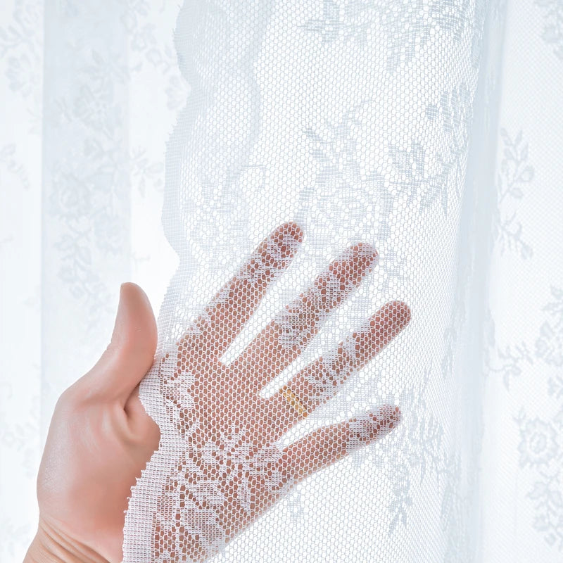 A hand is holding a sheer white lace curtain panel with floral embroidery.