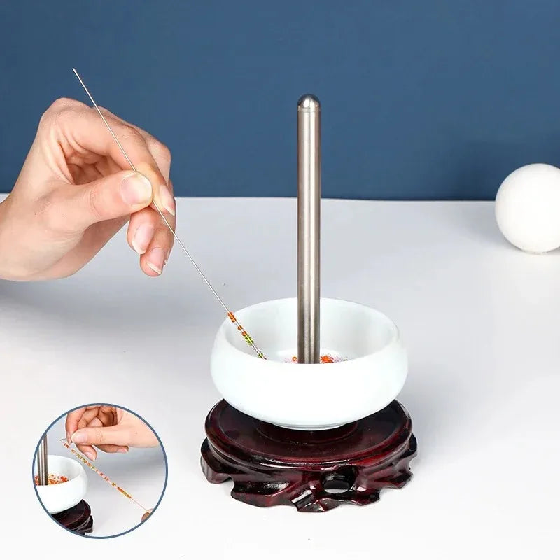 A hand is using a stainless steel beading needle to thread beads into a white bowl on a dark wooden stand.