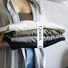 A hand holds a stack of folded white, olive green, and gray bed sheets secured with a white strap labeled Queen.