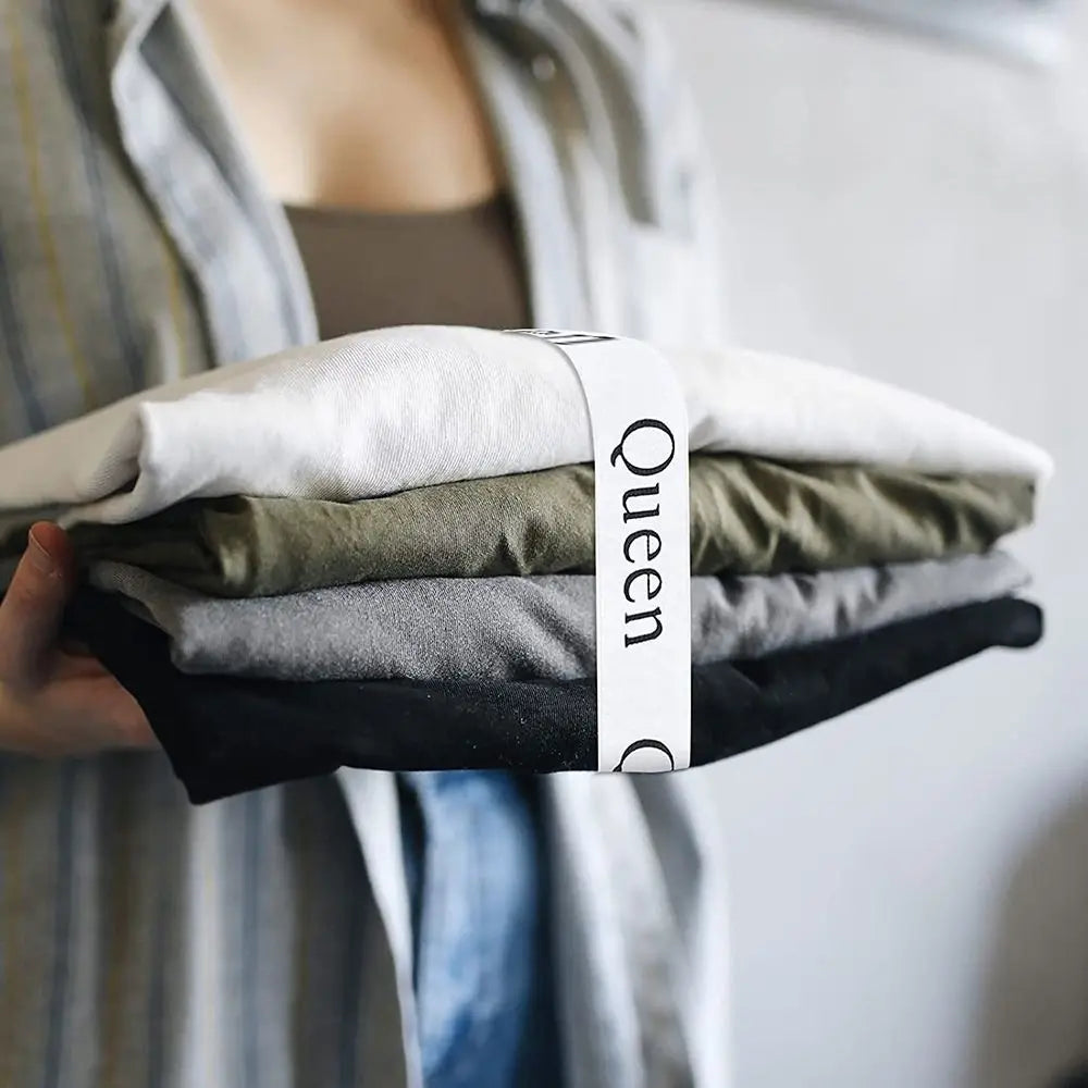 A hand holds a stack of folded white, olive green, and gray bed sheets secured with a white strap labeled Queen.