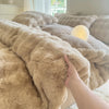 A hand is touching a fluffy beige rabbit fleece duvet cover on a bed.