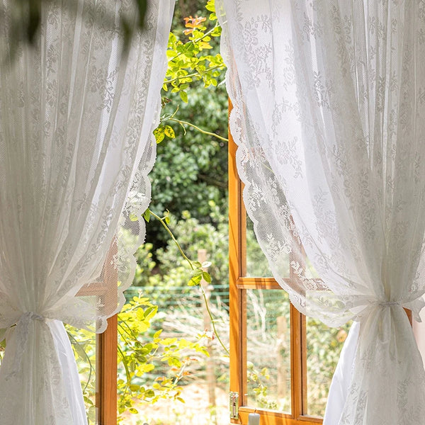 White sheer floral lace window curtains framing a view of green foliage outside