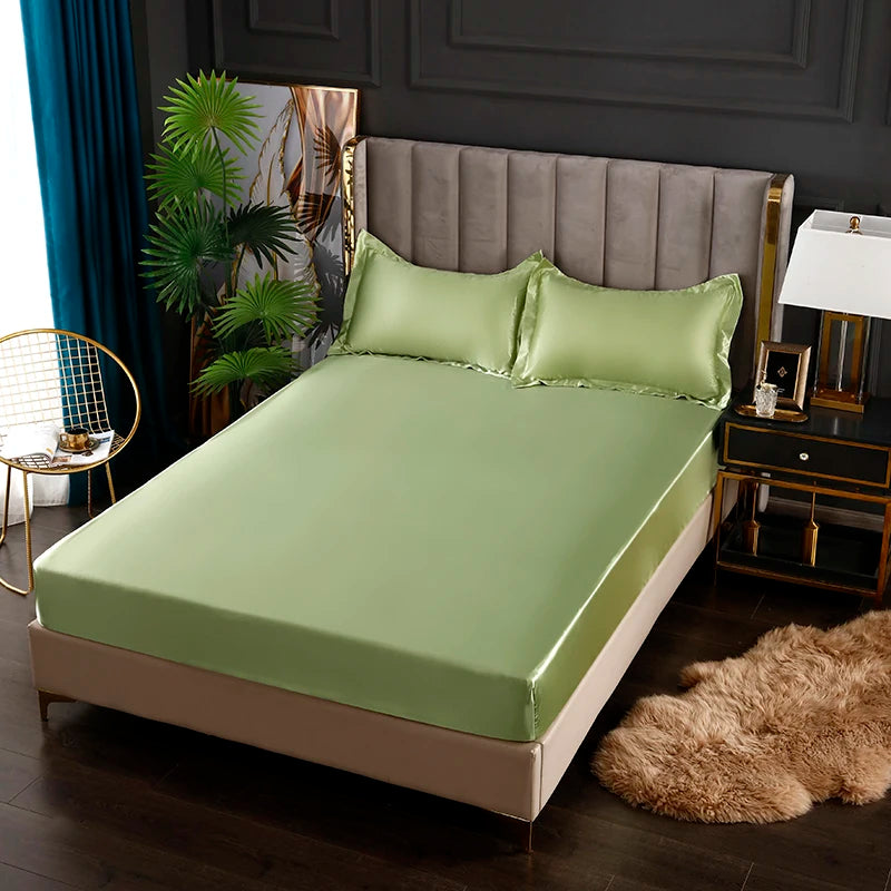 A light green satin fitted sheet is displayed on a bed with a gray upholstered headboard and dark wood accents.