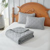 A set of light gray quilted pillows and a matching quilted bedspread displayed on a white bed with a wooden headboard.