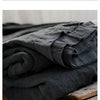 A stack of dark gray pure linen flat sheets resting on wooden crates and a bed.