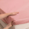 A hand gently touches a pink waterproof fitted bed sheet on a white mattress.
