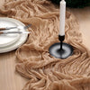 A beige crinkled cheesecloth table runner draped across a wooden surface with a black candle holder and white plates.