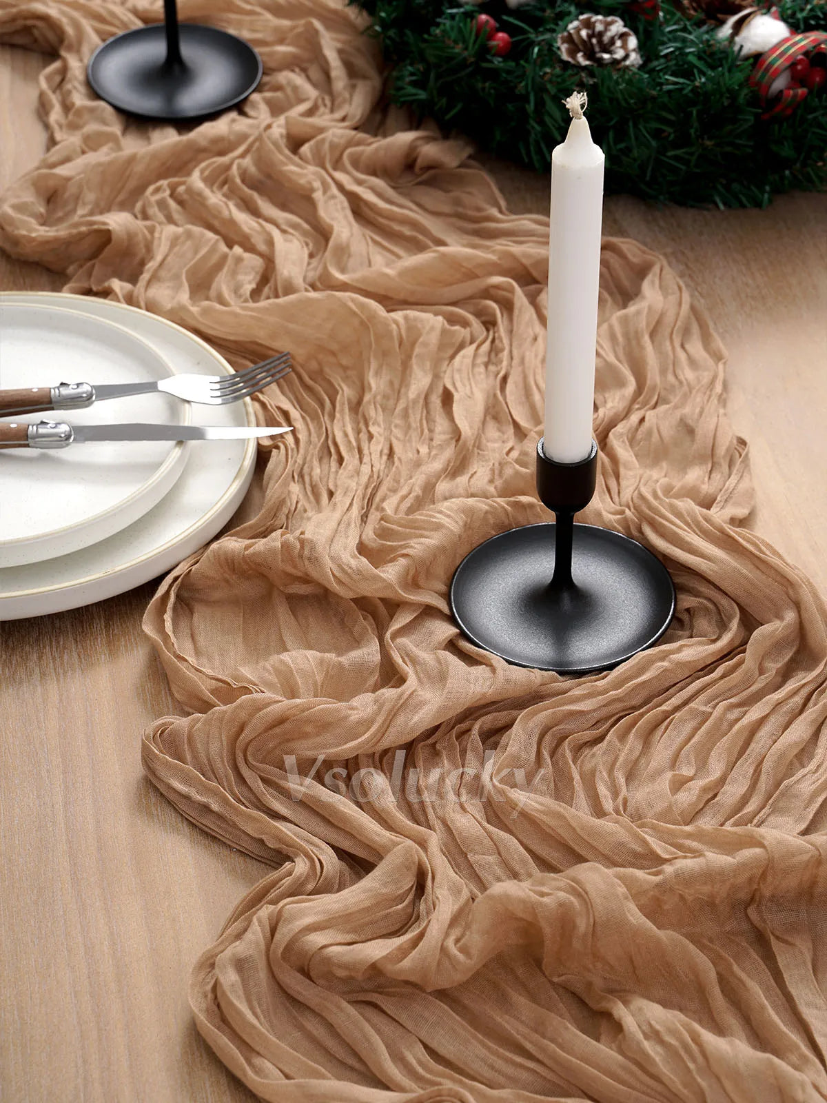 A beige crinkled cheesecloth table runner draped across a wooden surface with a black candle holder and white plates.
