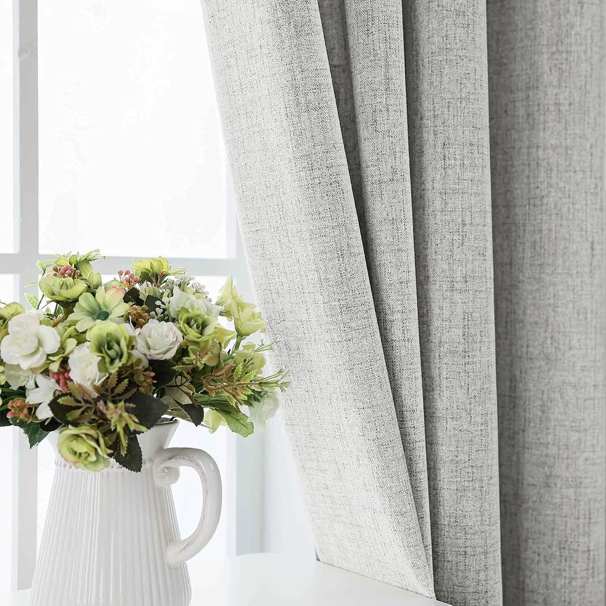 A white ceramic vase holds a bouquet of white and green flowers next to a gray linen curtain.