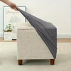 A hand is draping a gray jacquard stretch ottoman slipcover over a beige upholstered ottoman.