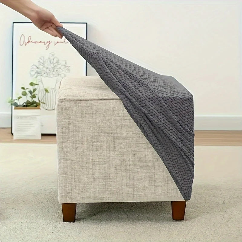 A hand is draping a gray jacquard stretch ottoman slipcover over a beige upholstered ottoman.