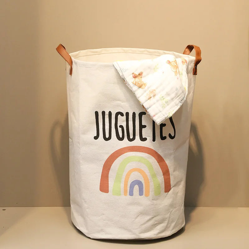 A white storage basket with a rainbow design and the word JUGUETES, featuring brown leather handles.