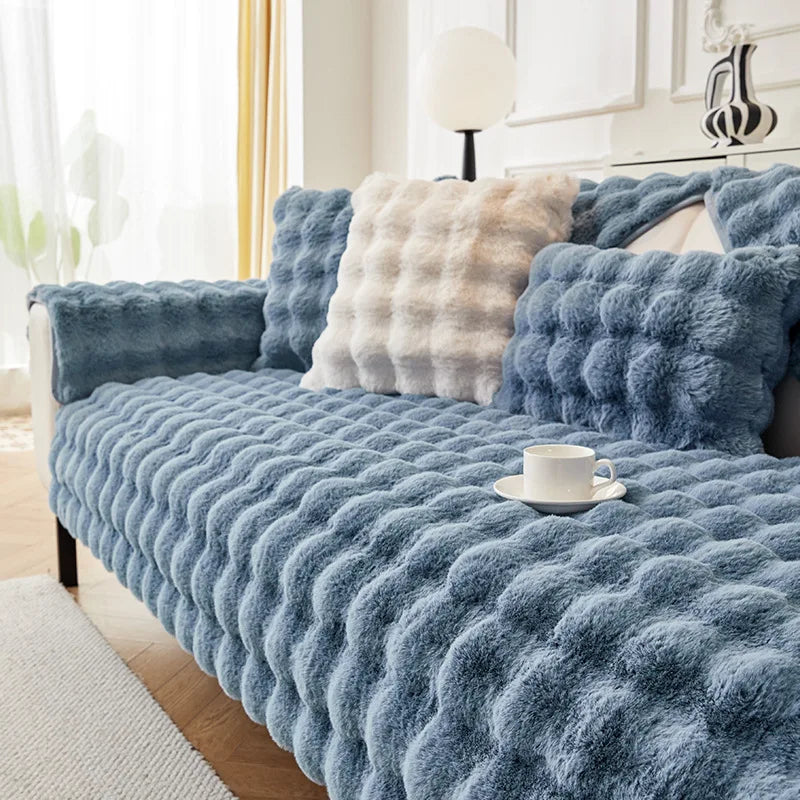 A blue plush rabbit fur sofa cushion cover with a cream textured pillow on a light-colored sofa.