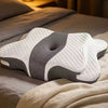 A white and gray ergonomic memory foam neck pillow resting on a bed with soft bedding.