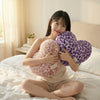 A young woman is sitting on a bed with two leopard print plush pillows, one pink and one purple.