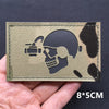 A rectangular tactical patch featuring a silhouette of a skull and crossbones with a camera, set against a khaki fabric backg