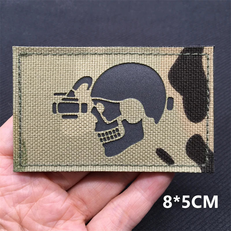 A rectangular tactical patch featuring a silhouette of a skull and crossbones with a camera, set against a khaki fabric backg