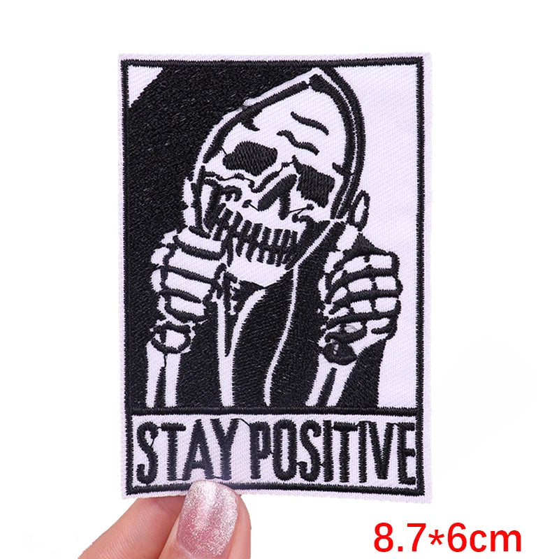 Black embroidered patch featuring a skull with hands covering its face and the text STAY POSITIVE
