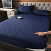 A dark blue quilted mattress protector is shown on a bed, with pillows and a bedside lamp visible.